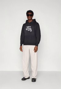 Sweatshirt com capuz preta com bolso frontal e design gráfico branco, combinada com calças largas bege e sapatos pretos sobre um fundo liso.