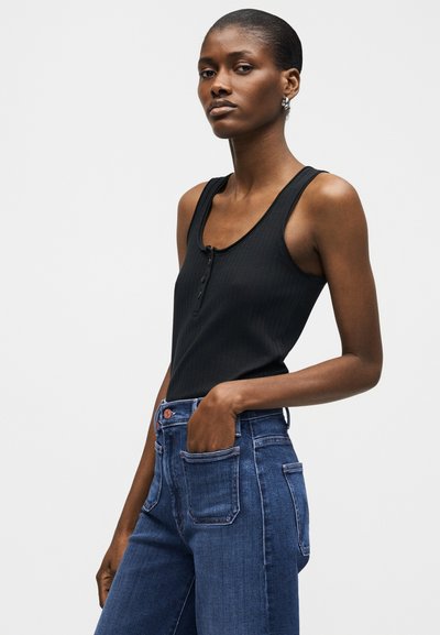 Γυναίκα με κοντά μαλλιά, που φοράει μαύρο ριμπ τοπ χωρίς μανίκια και μπλε τζιν ψηλόμεσο, στέκεται με το ένα χέρι στην τσέπη της.