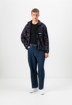 Young person standing, wearing black and purple patterned zip-up jacket, black shirt, loose blue jeans, and white sneakers against white background.