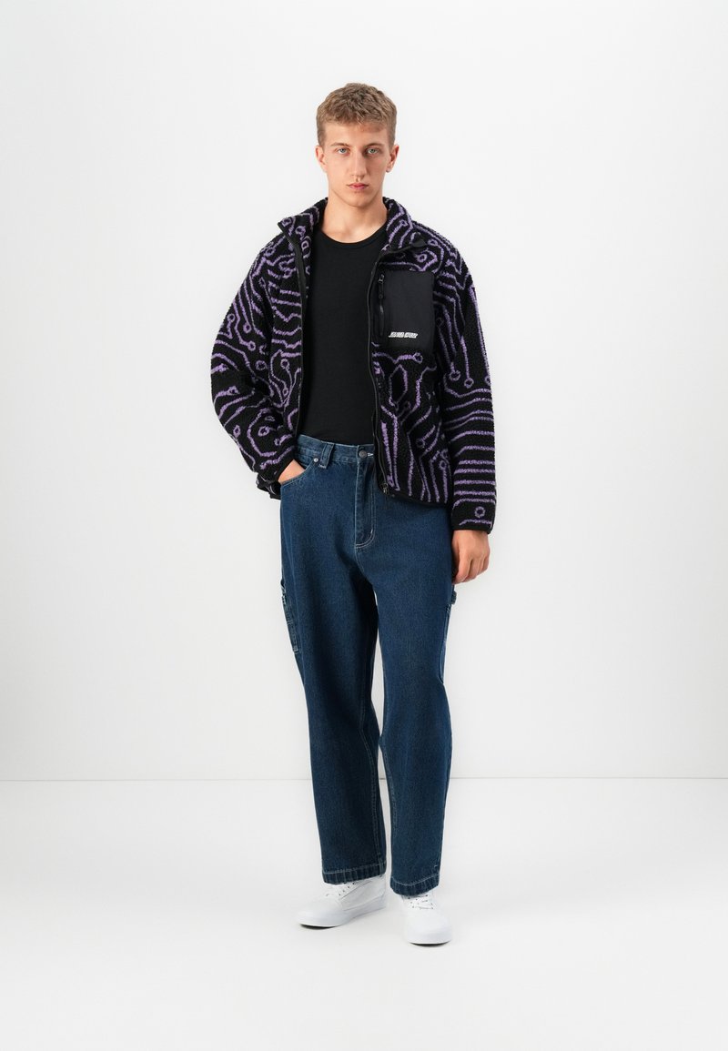 Jauns cilvēks stāv, ģērbies melnā un purpursarkanā rakstainā rāvējslēdzēja jakā, melnā krekla, brīvās zilajās džinsās un baltos sporta apavos pret balto fona.