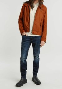 Veste légère marron avec fermeture éclair à l'avant, associée à un polo beige, un jean bleu foncé et des bottines noires. La veste présente un col à pression.