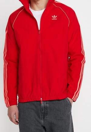 Red zip-up jacket made of lightweight fabric, featuring white Adidas logo and three white stripes along the sleeves. Textured finish.