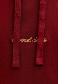 Bordeaux huppari, jossa on tekstuurikangas ja kultainen brodeerattu logo. Kaksi yhteensopivaa narua näkyy kaula-aukossa.