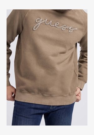 Sweat-shirt beige avec logo "guess" texturé en forme de corde sur la poitrine, porté par une personne les mains sur les hanches, associé à un jean bleu foncé.