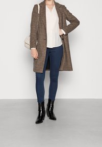 Brun houndstooth-lang frakke over en hvid bluse, parret med mørke skinny jeans og sorte hælede ankelstøvler, mens hun holder en cremefarvet quiltet håndtaske.