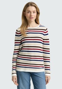 Vrouw met een lang mouw ribbeltrui met horizontale strepen in rood, blauw, bruin en beige, gecombineerd met blauwe spijkerbroek.