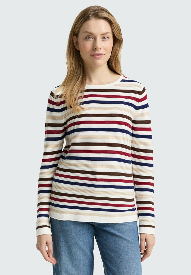 Vrouw met een lang mouw ribbeltrui met horizontale strepen in rood, blauw, bruin en beige, gecombineerd met blauwe spijkerbroek.