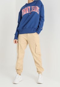 Blå sweatshirt med "TOMMY JEANS"-logotyp i rött, ihopparad medbeige cargobyxor med sidofickor och ribbade muddar, kompletterad med vita sneakers.