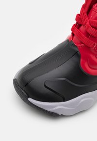 Czarno-czerwony but sportowy z gładką, syntetyczną cholewką, teksturowaną czarną podeszwą i gumowymi akcentami. Posiada czerwone sznurowadła i formowane kontury.