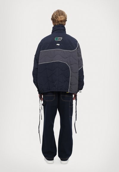 Puffer jacket in navy and gray with quilted design, white piping, and adjustable drawstrings at the waist. Features logo on the back.