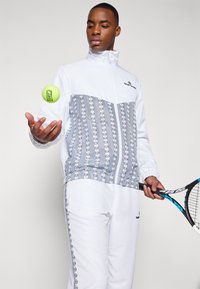 Beli športni trenirke z sivo patterned prsno ploščo, elastičnimi manžetami in detajlom logotipa, z žogico za tenis v roki in rekrom.