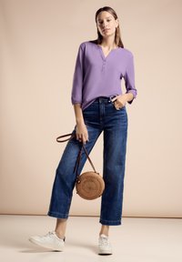 Maglietta lunga viola con scollatura henley, abbinata a jeans blu a vita alta. Il modello tiene una borsa rotonda di paglia e indossa sneakers bianche.