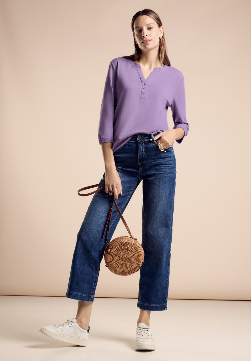 Maglietta lunga viola con scollatura henley, abbinata a jeans blu a vita alta. Il modello tiene una borsa rotonda di paglia e indossa sneakers bianche.