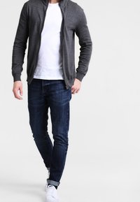 Szara bluza na zamek z kapturem na białym t-shircie, zestawiona z ciemnoniebieskimi jeansami i białymi trampkami. Casualowy strój o dopasowanym kroju.