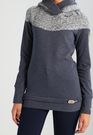 Bluza z kapturem z długim rękawem z melanżową szarą naramienną częścią i ciemnoniebieskim korpusem, noszona przez osobę w ciemnych spodniach na jednolitym tle.