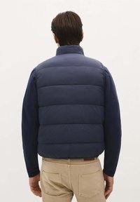Gilet imbottito blu navy con colletto alto, texture a rombi e senza chiusure visibili. Indossato sopra una camicia a maniche lunghe e pantaloni beige.