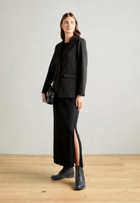 Veste noire avec deux poches avant, associée à une jupe noire avec des fentes sur les côtés. Complétée par des bottes noires à lacets sur un sol en bois.