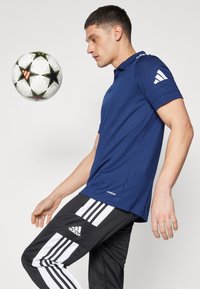 T-paita navy sinisessä värissä, jossa on teksturoitu pinta ja valkoisia yksityiskohtia, yhdistetty mustiin housuihin, joissa on kolme valkoista raitaa ja Adidas-logo. Jalkapallo leijuu lähellä.