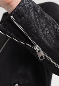 Svart jacka med texturerad tyg, silverfärgade dragkedjor och närbild av dragkedjedetaljerna vid ärmarna, som visar en jämn och glänsande yta.
