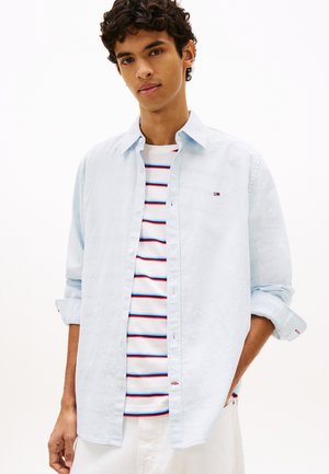 Joven con camisa azul claro abotonada sobre una camiseta blanca a rayas y pantalones blancos, de pie con una mano en el bolsillo.