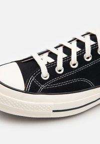 Svart lærret sneakers med hvite snørebånd, gummitåhette og kontrasterende hvite sømmer. Har metalløyer og teksturert såle.
