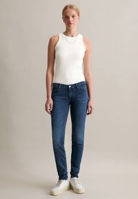 Top sans manches blanc, jeans slim-fit bleu foncé et baskets blanches. Les jeans ont des poches et un léger délavage sur les jambes. Accessoire en chaîne en argent simple.