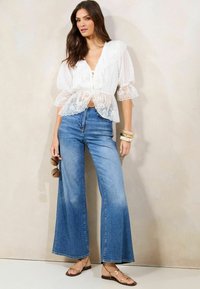 Blusa bianca in pizzo con maniche a sbuffo, abbinata a jeans in denim blu a vita alta e a zampa. La modella tiene degli occhiali da sole e indossa sandali color marrone chiaro.