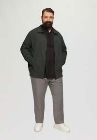Dunkelgrüne Zip-Jacke über einem schwarzen Shirt, kombiniert mit hellgrauen Hosen und weißen Sneakers. Das Outfit hat einen lässigen Schnitt und eine strukturierte Stoffbeschaffenheit.