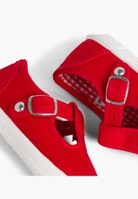 Zapatillas de lona rojas con una suela de goma blanca, que presentan una hebilla plateada y un diseño recortado, forradas con un patrón de cuadros rojo y blanco.