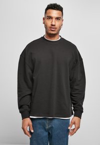 Hombre con cabello corto y rizado y arete, vistiendo un sudadera negra de gran tamaño sobre una camisa blanca y jeans azules, de pie contra un fondo gris.