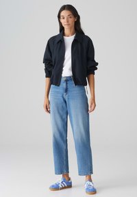 Veste noire zippée avec des poignets élastiques, portée par-dessus un t-shirt blanc. Jeans droit bleu clair et chaussures de sport bleues avec des accents blancs.