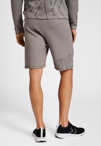 Grå shorts i mjukt material med en ren design, prydda med en svart geometrisk logotyp på underbenet. Kombinerat med svarta träningsskor.