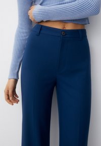 Pantalon bleu marine ajusté avec une texture lisse, doté d'une fermeture à bouton, de poches latérales et d'une coupe droite.