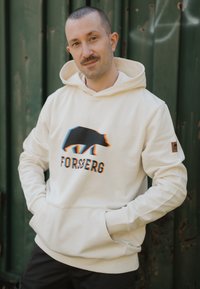 Sudadera color crema con un gráfico de un oso en negro y acentos de arcoíris, con un bolsillo frontal y puños acanalados. Parche del logo en la manga.
