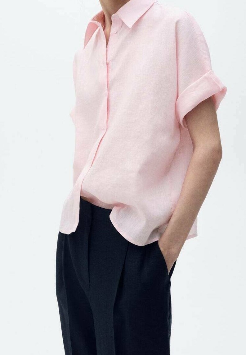 Personne portant une chemise à manches courtes rose clair boutonnée avec les manches retroussées et un pantalon noir taille haute, main dans la poche.