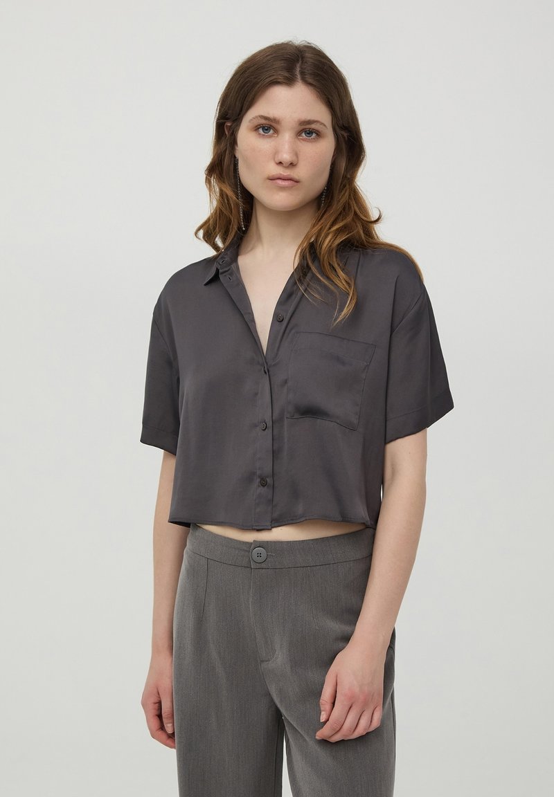 Camicia corta a maniche corte in tessuto satinato grigio scuro. Presenta un colletto, chiusura a bottoni e una tasca sul petto. Abbinata a pantaloni grigi sartoriali.