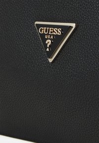 Sort struktureret læder håndtaske med et guld og sort trekantet logo-plade med "GUESS USA" og et spørgsmålstegn-design.