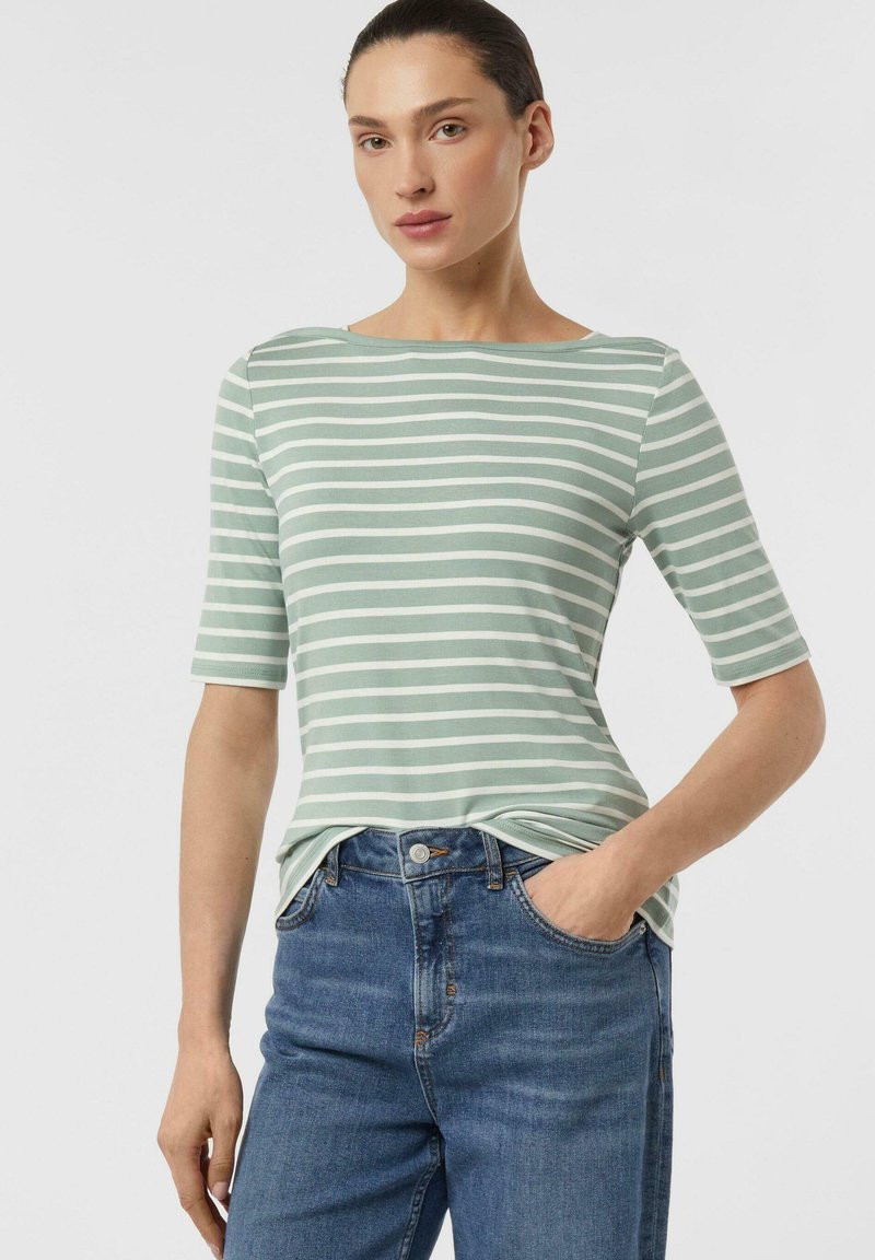 Frau trägt ein grün-weiß gestreiftes kurzärmeliges Oberteil mit Jeans, eine Hand in der Tasche, steht vor einem einfachen weißen Hintergrund.