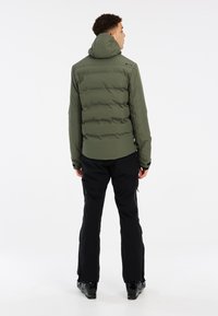 Chaqueta acolchada verde oliva con capucha, que presenta una textura acolchada. Se lleva con pantalones de nieve negros y botas de esquí negras. Tiene un diseño ajustado.