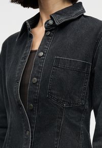 Κοντινό πλάνο ενός ατόμου που φοράει μαύρο πουκάμισο από τζιν με κουμπιά και τσέπη στο στήθος πάνω από ένα σκούρο ριγέ τοπ.