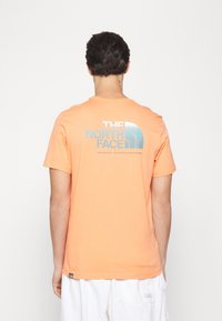 Pomarańczowa bawełniana koszulka z dużym niebieskim logo na plecach, okrągły dekolt i krótkie rękawy; gładka faktura, luźny krój.