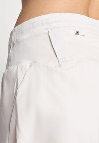 Detail osoby nosiacej biele nohavice s elastickým látkovým pásom a malým zadným vreckom na zips.