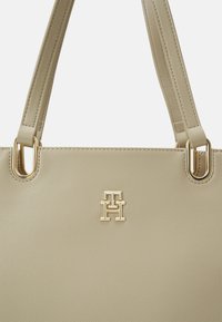 Sac à main texturé beige avec quincaillerie dorée. Présente des poignées doubles et un logo doré proéminent. Surface lisse avec une forme structurée.