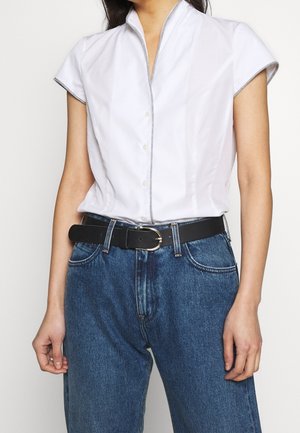 Nainen, jolla on valkoinen lyhythihainen paita, joka on puettu sinisiin farkkuihin ja musta vyö hopeisella soljella.