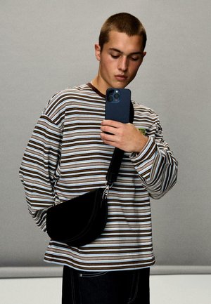 Svart axelremsväska med en böjd form, slät textur; bärs över en flerfärgad randig långärmad skjorta. Smartphone hålls i handen.