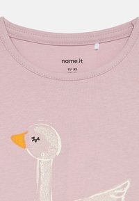 Lys rosa bomull T-skjorte med rund hals. Har et hvitt andemotiv med oransje nebb og svarte øye detaljer. Merket "name it."