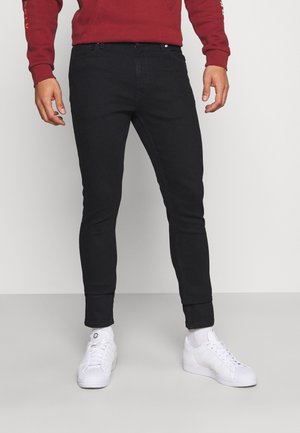Mann trägt schwarze Slim-Fit-Jeans, weiße Turnschuhe, weiße Socken und ein rotes Langarmshirt und steht vor einem schlichten Hintergrund.