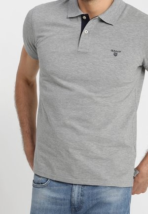 Mężczyzna w szarej koszulce polo z krótkim rękawem z czarną lamówką przy listwie i jasnoniebieskich dżinsach, stoi z rękami w kieszeniach.