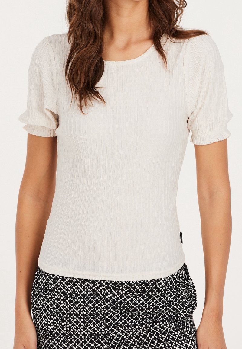 Femme portant un haut texturé à manches courtes de couleur écrue associé à un pantalon à motifs noirs et blancs, devant un fond neutre.