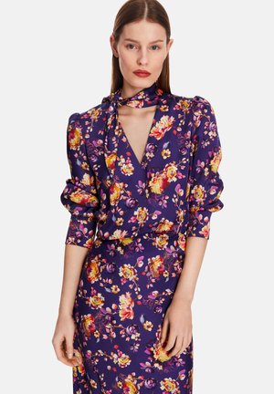 Blumen-Wickeloberteil aus lila Stoff, mit kräftigen gelben und pinken Blumen, langen Ärmeln und einer Nackenbindedetail. Glatte Textur, körperbetonter Schnitt.